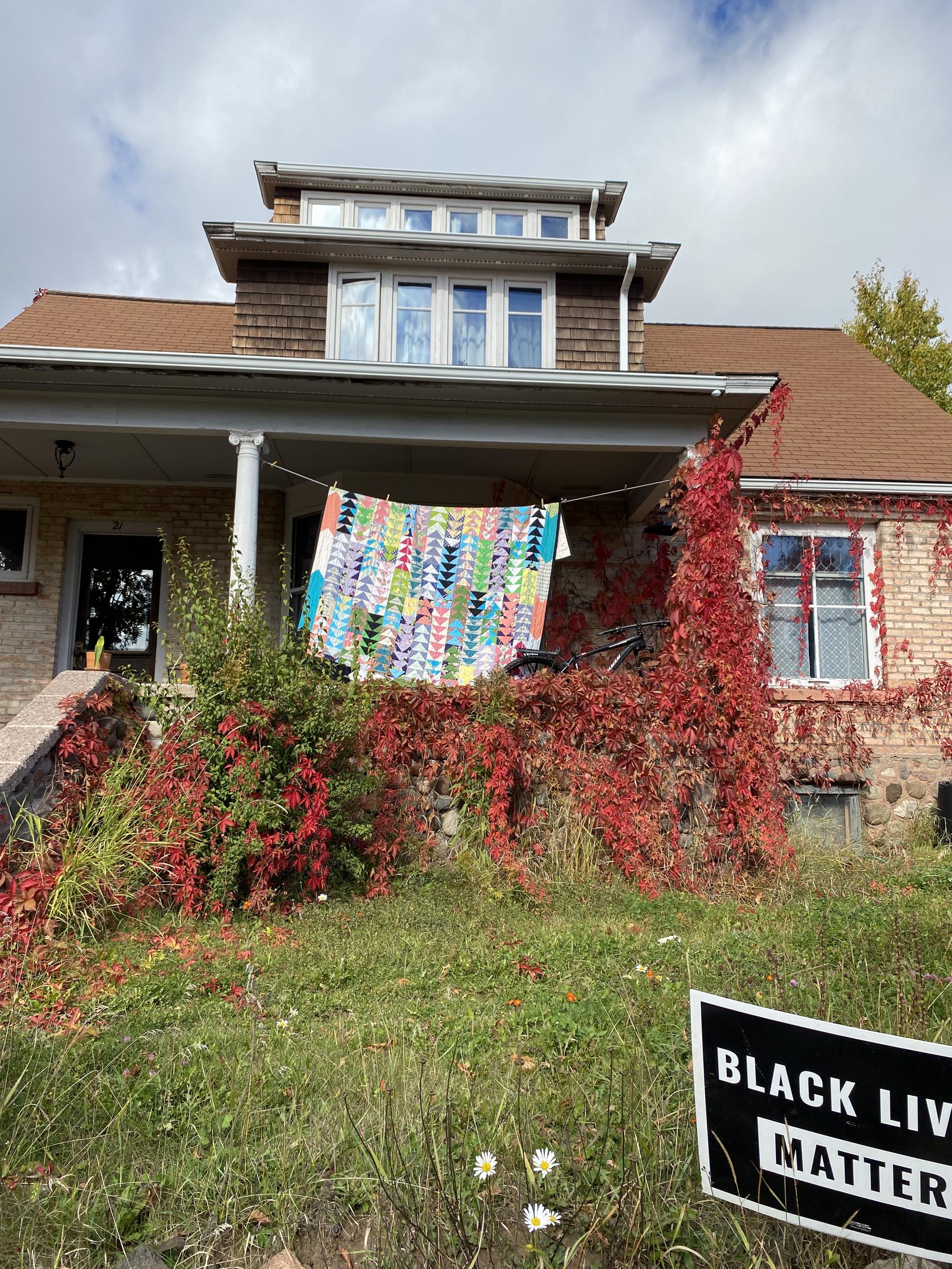 photo of the front porch of a house with a vibrant geometric quilt hanging from a clothesline, outshining the fall foliage climbing up the walls