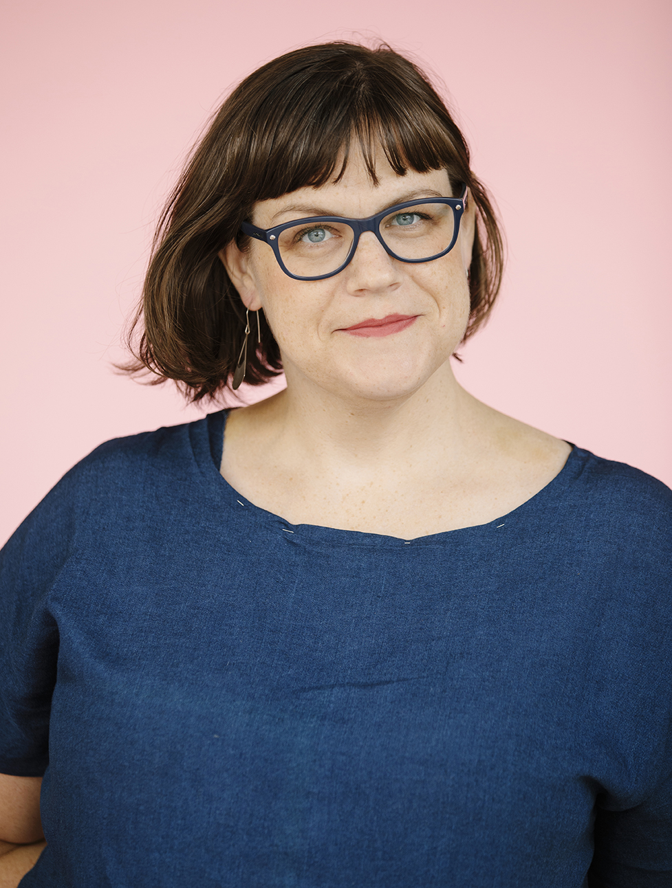 studio portrait of Laura Brown, a white woman with brown hair wearing a blue dress and dark-rimmed glasses faces the camera